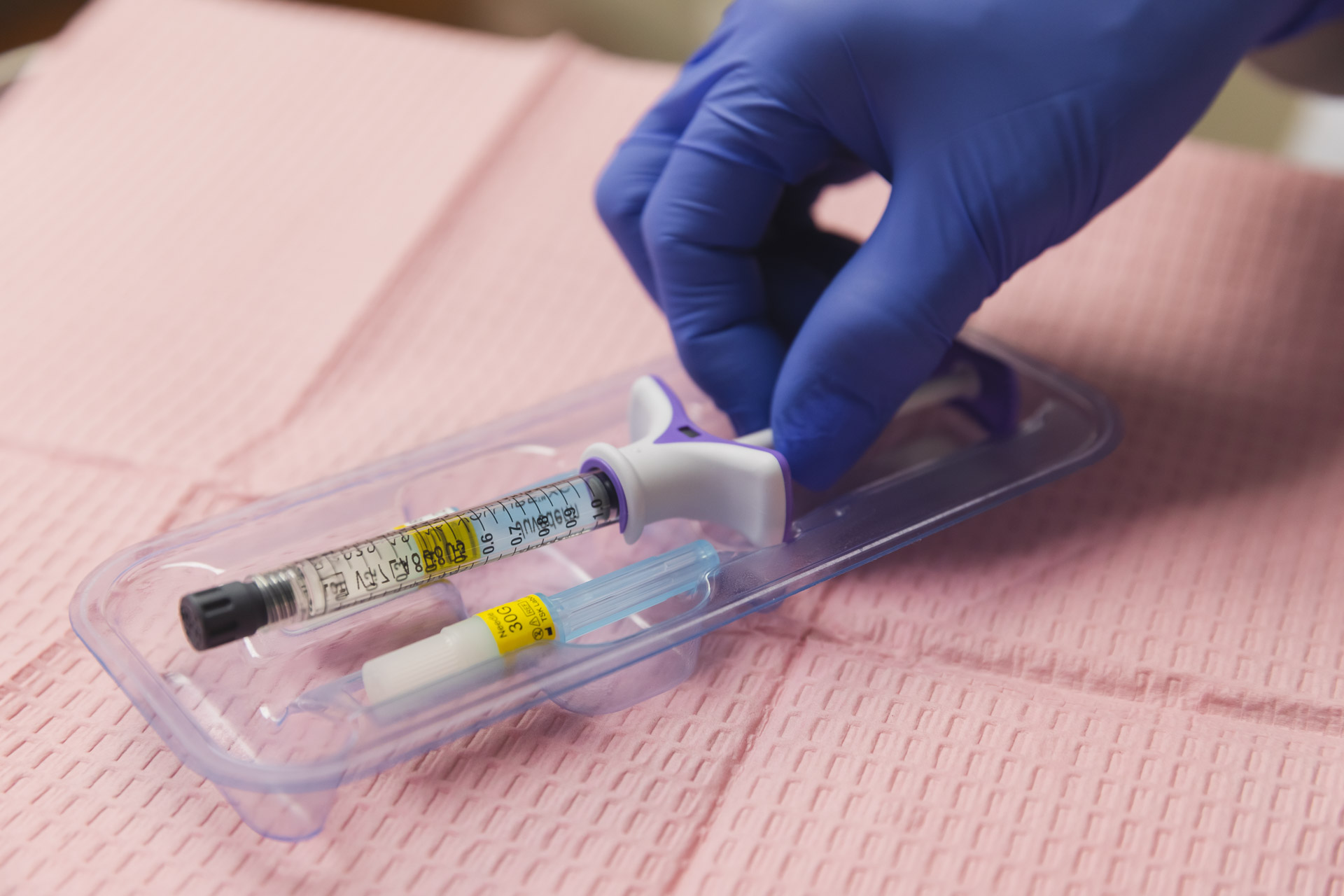 The hand of a provider, wearing a surgical glove, reaches for a pre-measured syringe of dermal filler. Syringes like these are used during lip fillers in Arlington to enhance the shape and volume of the lip.