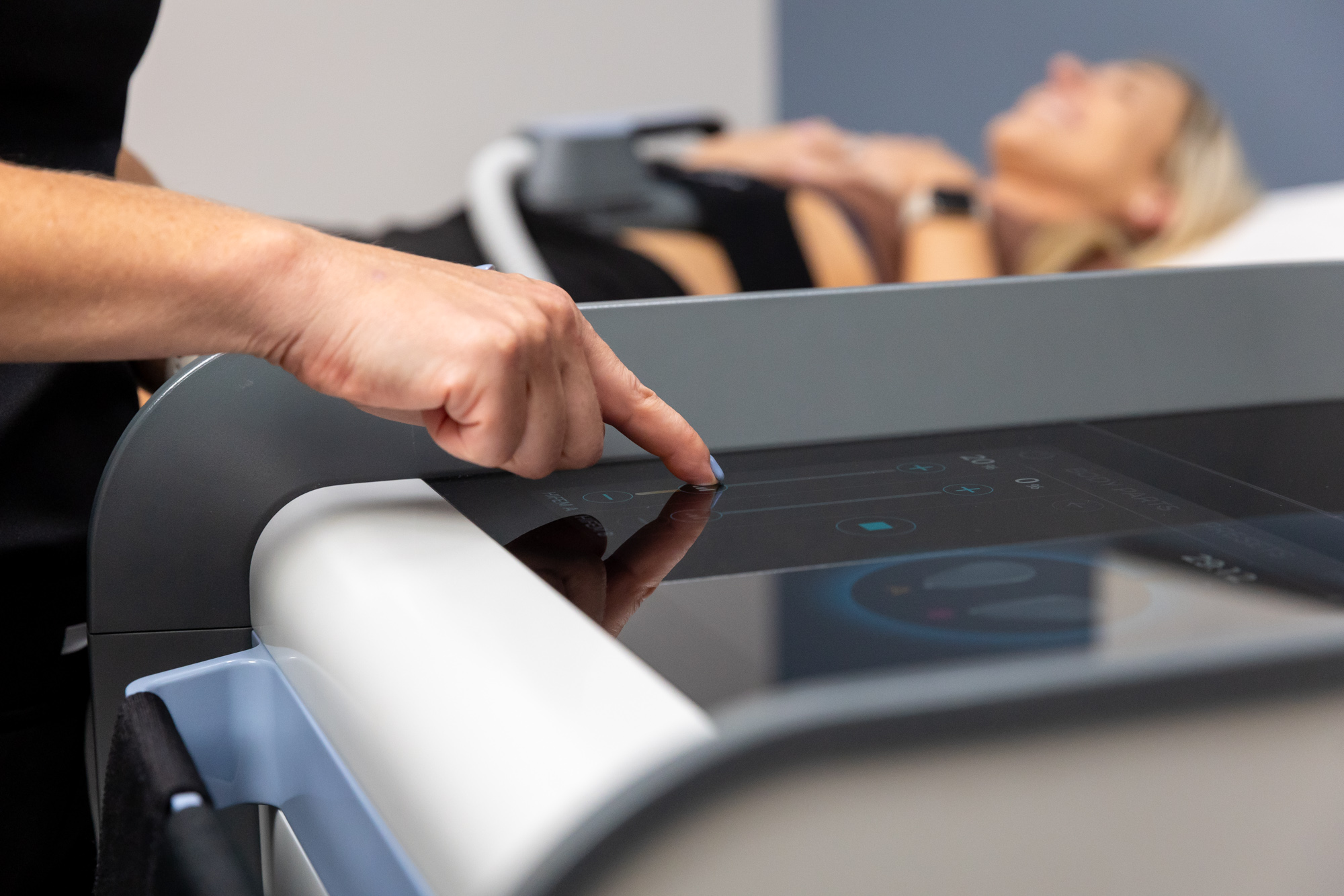  A provider at the Institute for Hormonal Balance slides the controls on the screen of the Emsculpt NEO device. A patient lies behind her receiving Emsculpt body sculpting in Arlington.